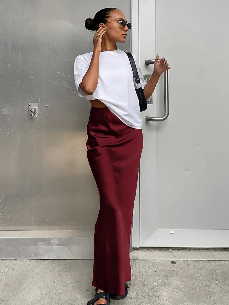 Woman wearing a white top and maroon pants standing against a gray wall.