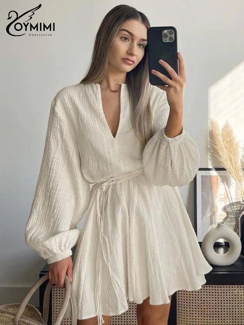 Woman taking a mirror selfie wearing a white dress with long sleeves, holding a phone.