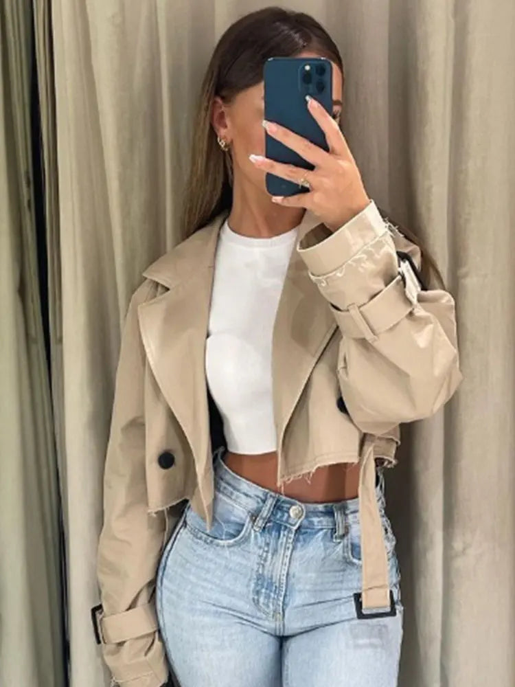 Person wearing a beige trench coat over a white top and blue jeans, taking a mirror selfie.
