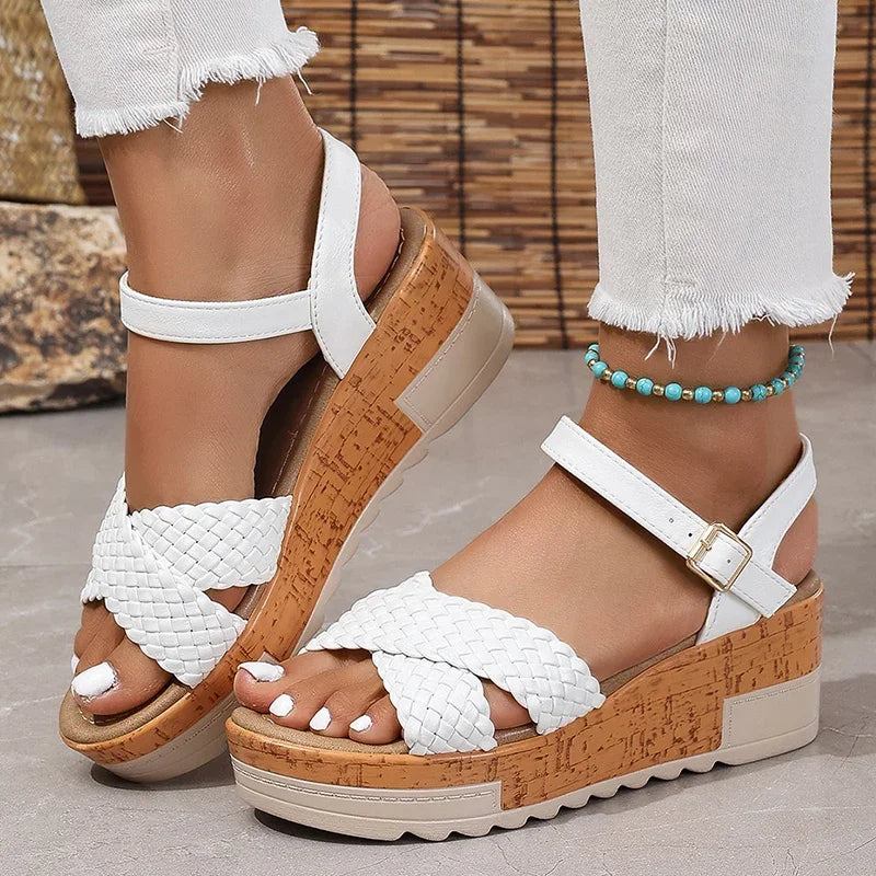 White sandals with brown soles worn by a person with white pants and a bracelet.