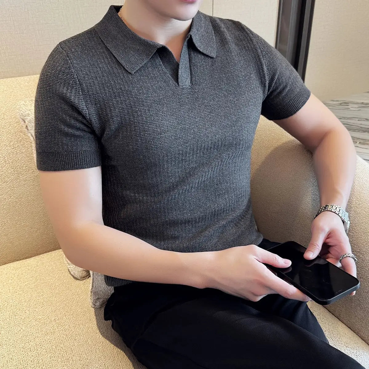 Person wearing a gray polo shirt sitting on a beige couch holding a phone.