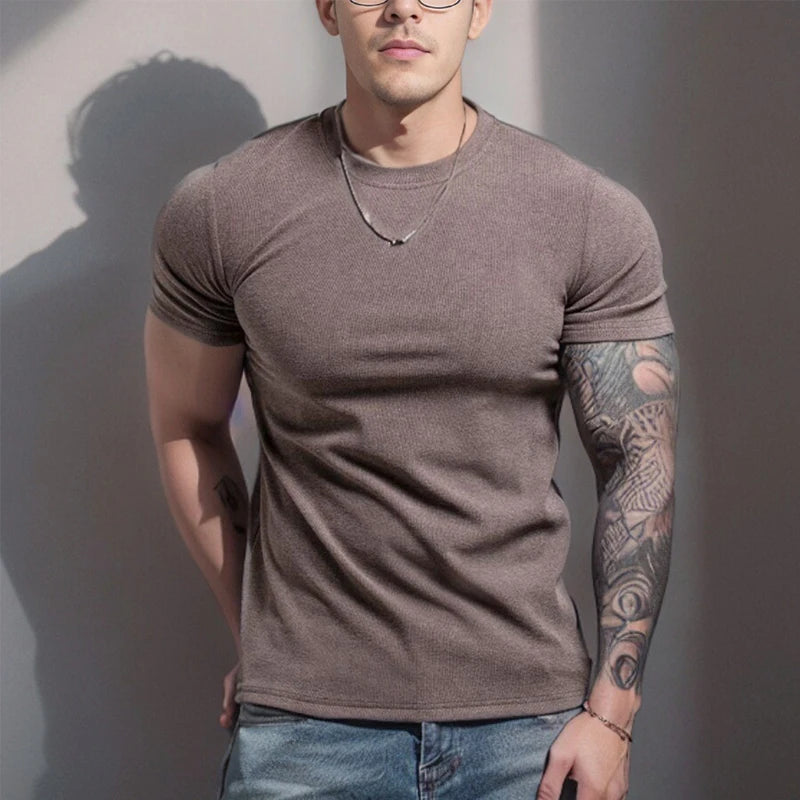 Man wearing a brown t-shirt and blue jeans with tattoos on arms against a plain background
