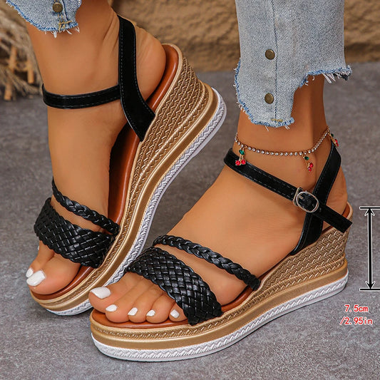 Black wedge sandals with braided straps worn by a person on a gray surface.