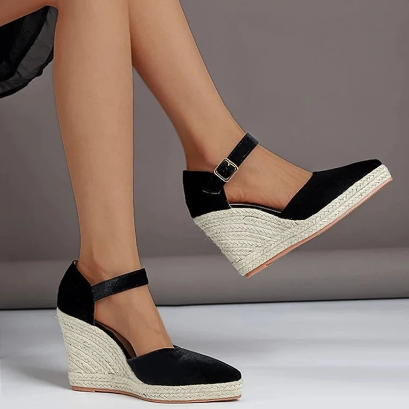 Black wedge sandals with espadrille sole worn by a person on a gray background