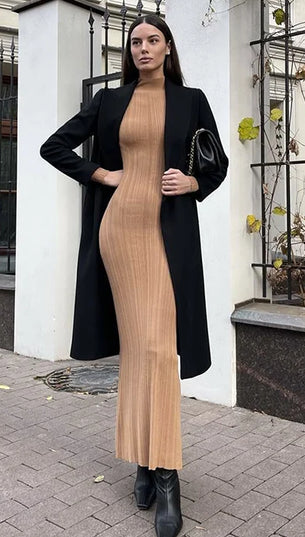 Woman in a long black coat and beige dress standing on a street with buildings in the background.