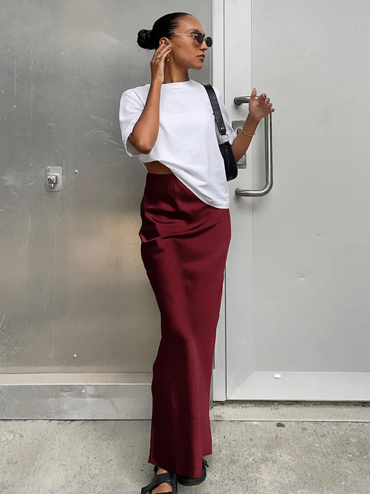 Woman wearing a white top and maroon pants standing against a gray wall.