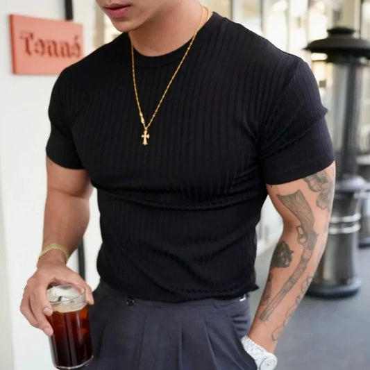 Person wearing a black t-shirt and gold necklace, holding a drink in an indoor setting.