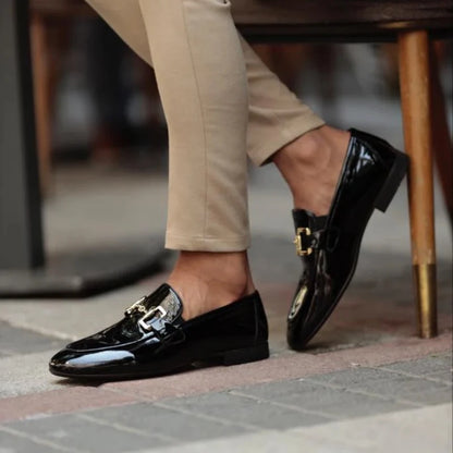 Black patent leather loafers worn with beige pants on a blurred background