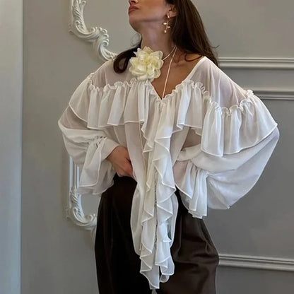 Woman wearing a white ruffled blouse with a decorative flower accessory against a neutral wall.