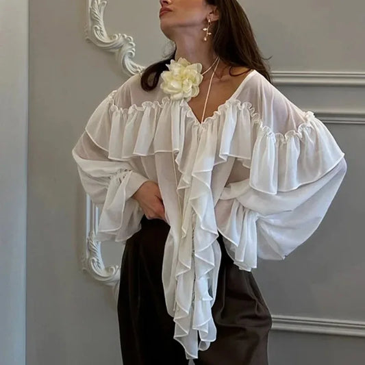 Woman wearing a white ruffled blouse with a decorative flower accessory against a neutral wall.