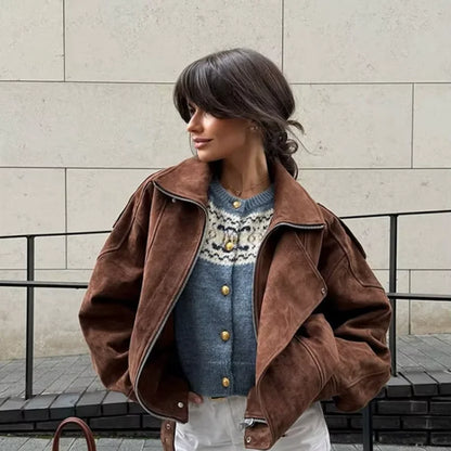 Woman wearing a brown suede jacket over a patterned sweater against a beige wall.