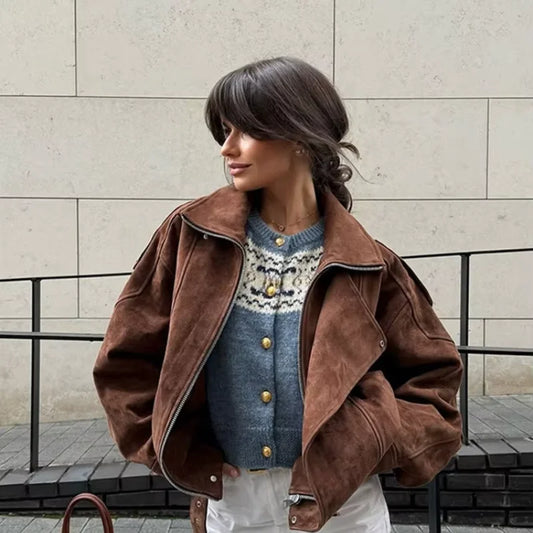 Woman wearing a brown suede jacket over a patterned sweater against a beige wall.