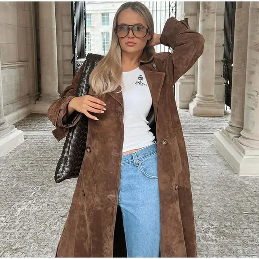 Woman wearing a brown coat, white shirt, and blue jeans in an urban setting.