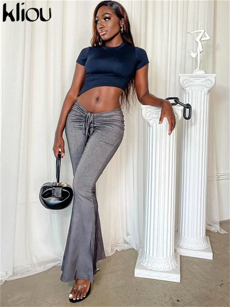 Woman in navy top and gray flared pants holding a black handbag against a white curtain background.