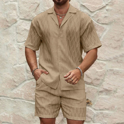 Man wearing a beige short-sleeve shirt and shorts set against a stone wall.