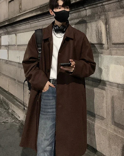 Person wearing a brown coat and face mask standing against a stone wall.
