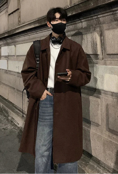 Person wearing a brown coat and face mask standing against a stone wall.