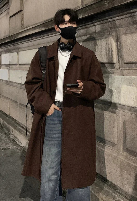 Person wearing a brown coat and face mask standing against a stone wall.