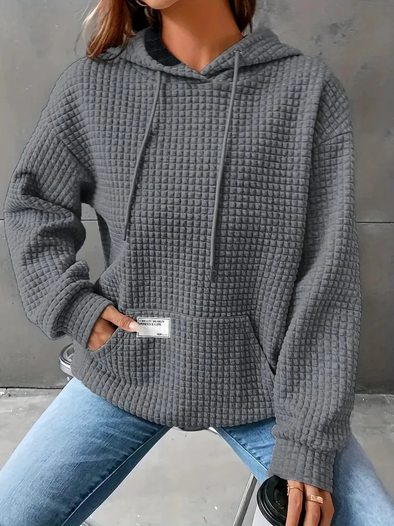 Person wearing a gray textured hoodie with a visible brand label, sitting against a neutral background.