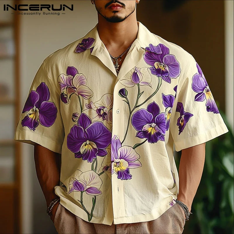 Man wearing a beige shirt with purple floral patterns, standing against a blurred indoor background.