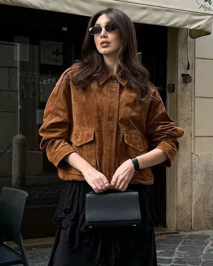 Woman in a brown jacket and sunglasses holding a black clutch in an urban setting.