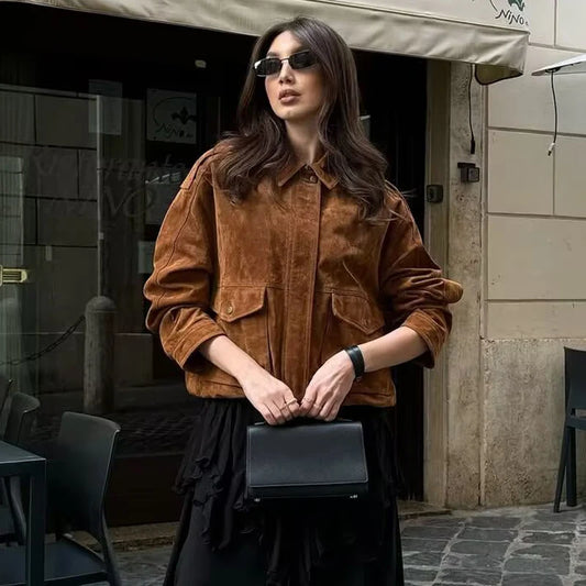 Woman in a brown jacket and sunglasses holding a black clutch in an urban setting.