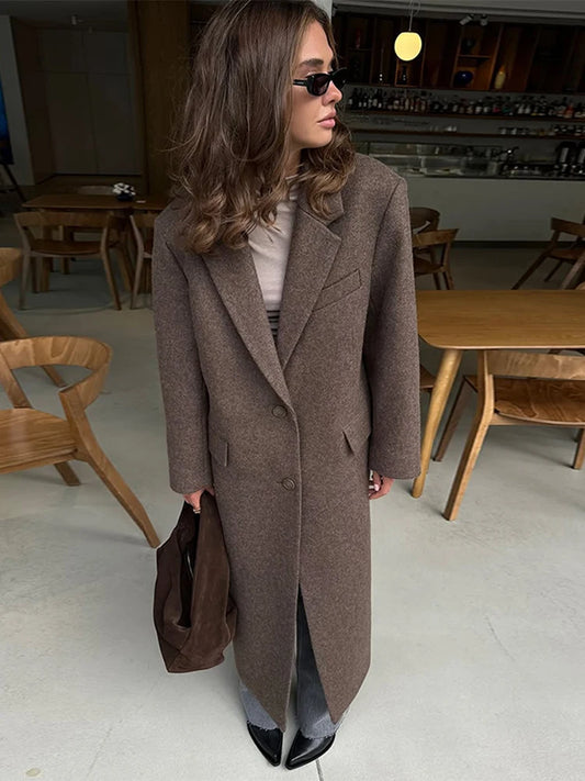 Person wearing a long brown coat in an indoor setting with wooden furniture.