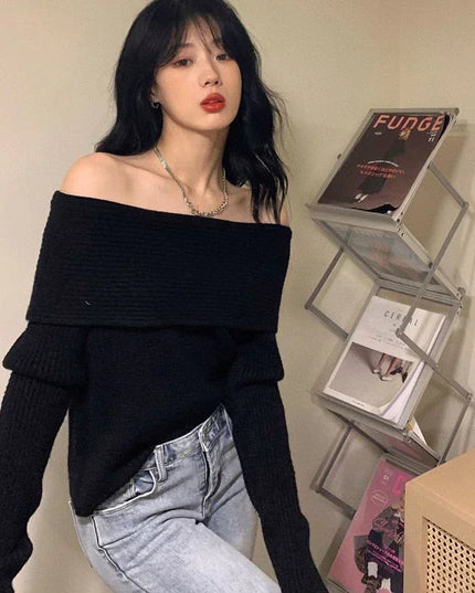 Woman wearing a black off-shoulder sweater and light blue jeans, standing in front of a shelf with books and decorative items.