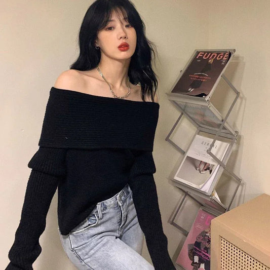 Woman wearing a black off-shoulder sweater and light blue jeans, standing in front of a shelf with books and decorative items.