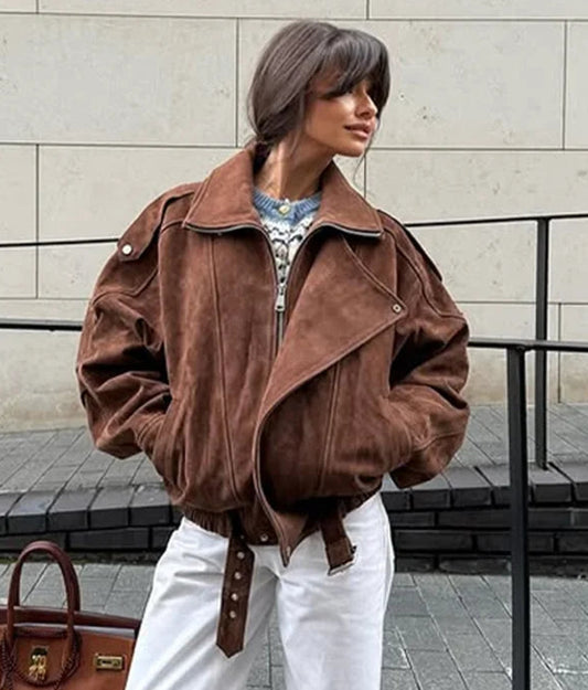 Person wearing a brown jacket and white pants with a brown handbag, standing against a stone wall.